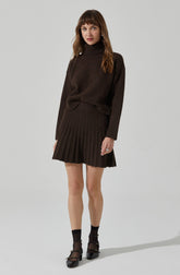 A woman is wearing a dark brown turtleneck sweater with long sleeves and a matching ribbed pleated skirt that falls above the knees. She is also wearing black ankle socks and black shoes with ankle straps.